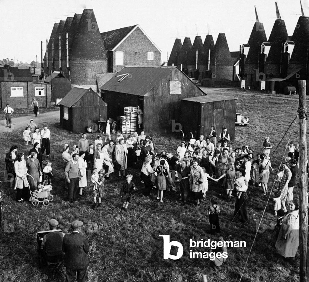 Hop pickers gathered in a Kent village with oast houses in the background, c. 1930 (b/w photo)