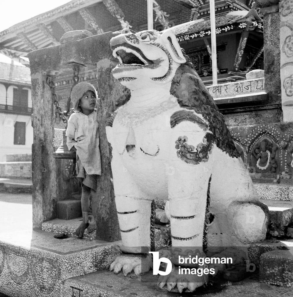A young boy lookes weary at a statue of a dragon dog guarding one of the buddhist temple in Katmandu, Nepal, February 1961 (b/w photo)
