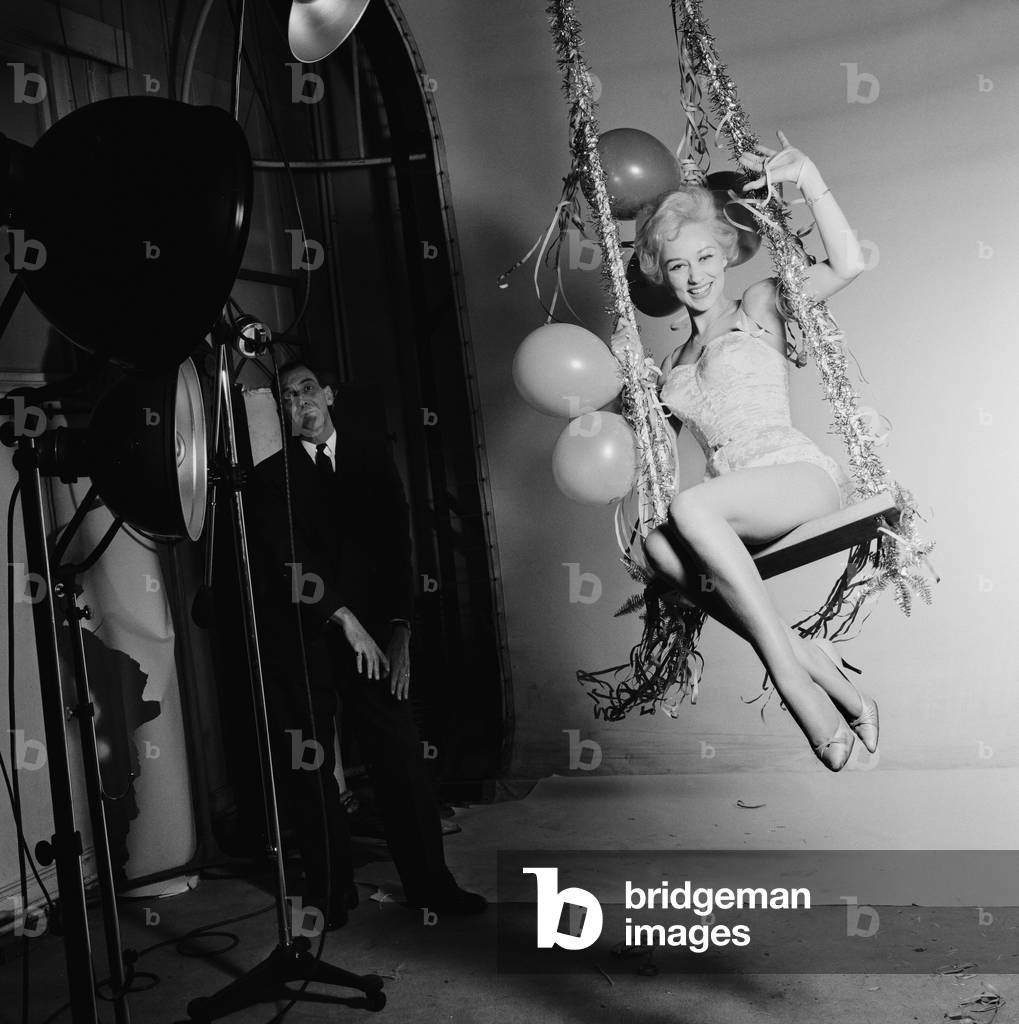 Actress Carole Leslie posing in the studio wearing a white dress, sitting on a swing with Christmas decorations.
24th December 1958.