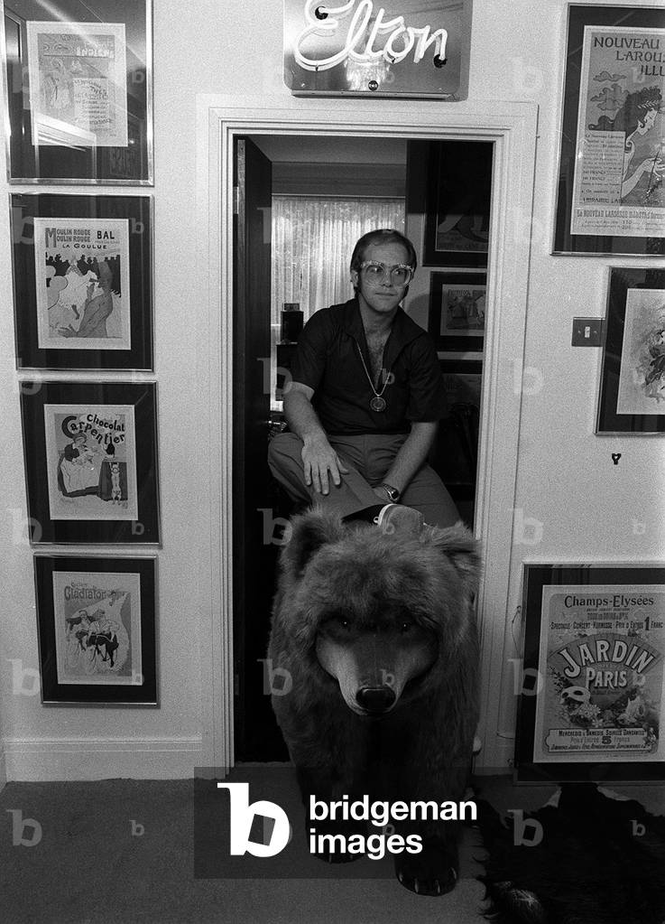 Sir Elton John in his new £50,000 mansion in Camberley, Surrey sitting on top of a bear June 1974 (b/w photo)