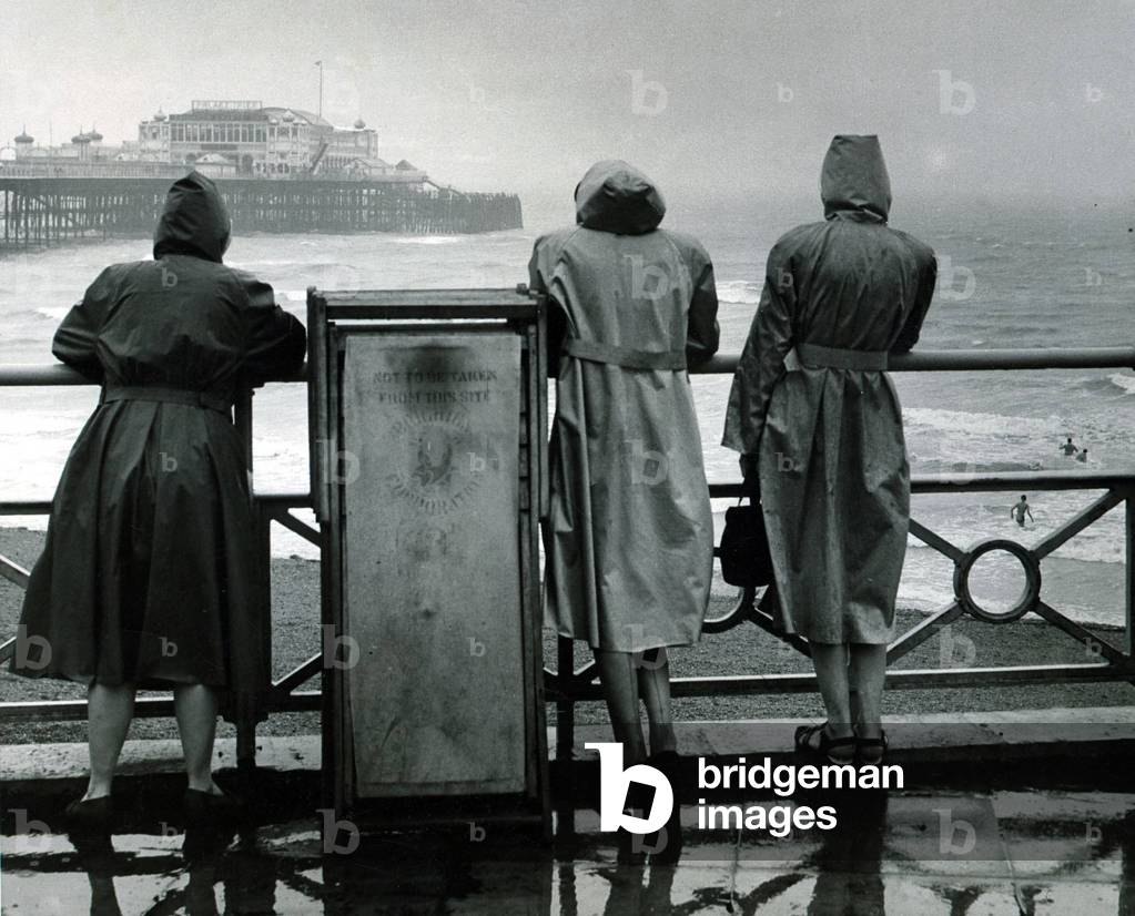 Three girls in raincoats watch members of the Brighton Swimming Club having a midday dip from a deserted beach, July 1949 (b/w photo)