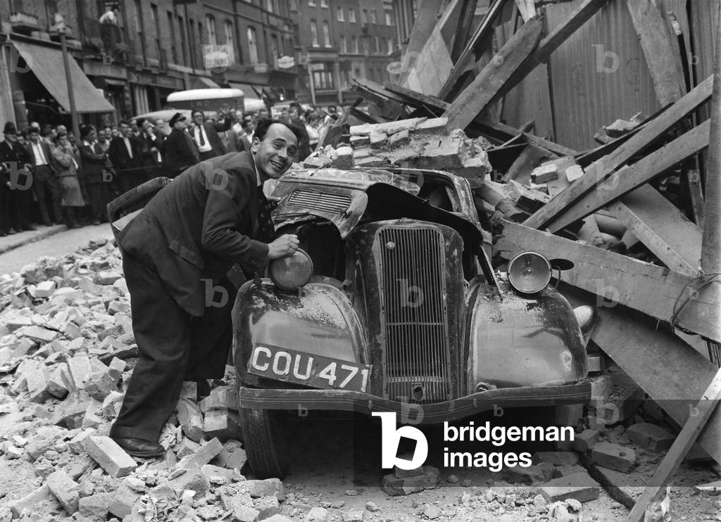 No wonder Hugh Walters, the man with the car in this picture is smiling. He's lucky to be alive
Lucky that he was not still sitting at the driving wheel when tons of rubble came crashing down in Lisle-street, Soho, yesterday ( and crushed his old saloon car. 
School teacher Walters, 27, left his home in Beatrice-avenue, Norbury, and drove into London to keep an appointment at a hospital in Soho. Friends had warned him: Be careful where you park your car. The police are always towing cars away. Hugh looked around carefully when he reached Lisle-street. There was a nice clear space near a hoarding. He parked there, nipped out and went to the hospital. Fifteen minutes later he returned - to Chaos. There were fire engines, ambulances, crowds and a pall of dust. A four- storey building under demolition had collapsed and fifty tons of brickwork lay in the roadway. Police had seen the danger and cleared the street of people in time. No one was hurt. The only casualty was Hugh's car. Hours later it was towed out, dented, smashed and bent. July 1960