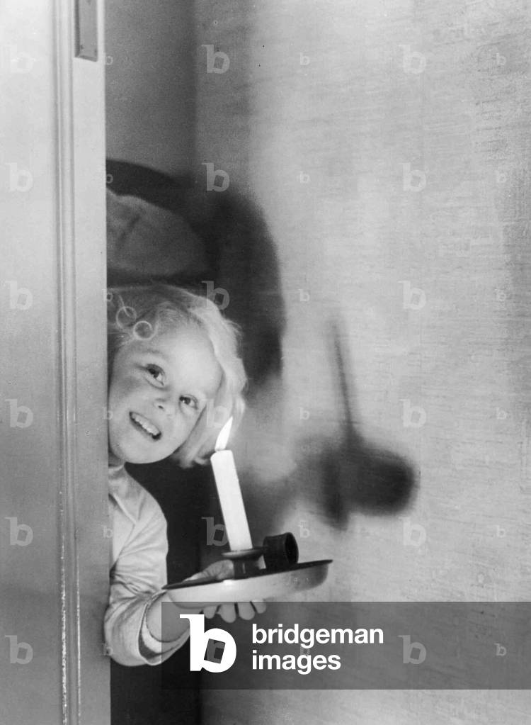 Young girl peering behind a door holding a candle, c.1945 (b/w photo)