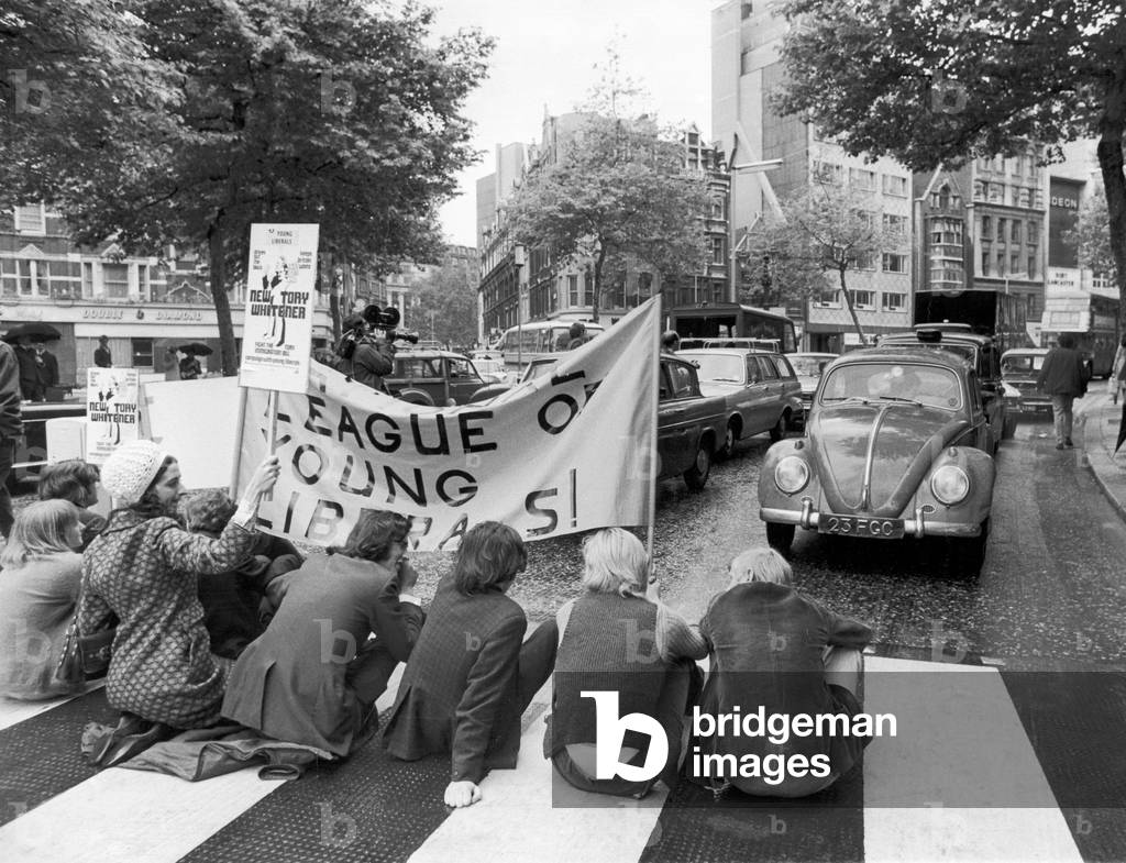 A group of young Liberals stop traffic by sitting on a pedestrian crossing in a busy London street in protest against the Government's Immigration Bill. Drivers of vehicles were asked to show passes by Ashley Wood who dressed in a South African policeman's uniform. 18th June 1971 (b/w photo)