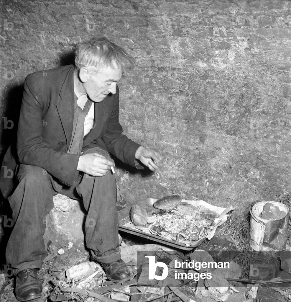One of two homeless gentlemen who live in the cellar of a bombed out building close to Tower Bridge, seen here gather lunch together, 1953 (b/w photo)
