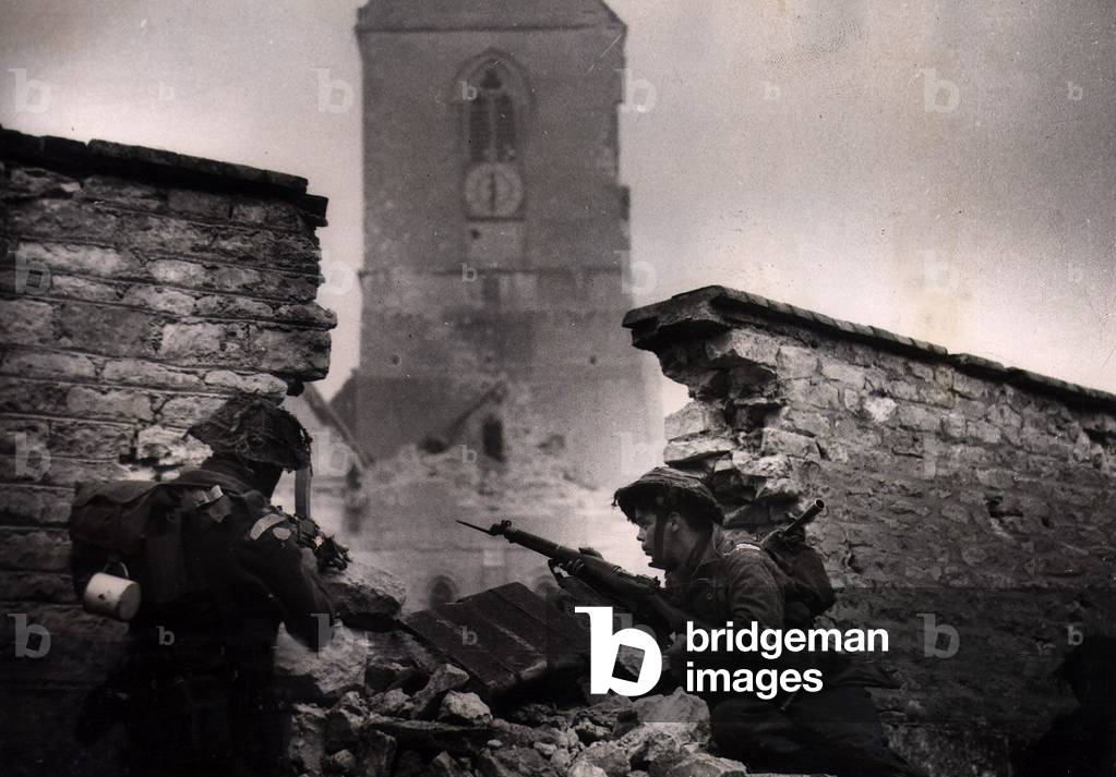 Soldiers from the British army seen here moving cautiously through the ruined wall in St Maugieu, June 1944 (b/w photo)