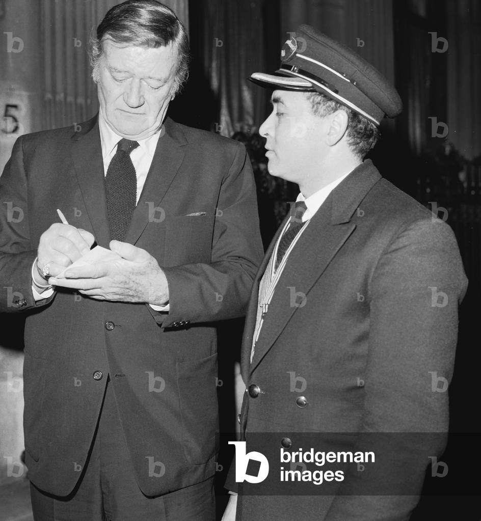 American actor John Wayne leaving the Ambassadeurs Club in Hamilton Place, London signs an autograph for doorman Tas Alder.
17th January 1974.