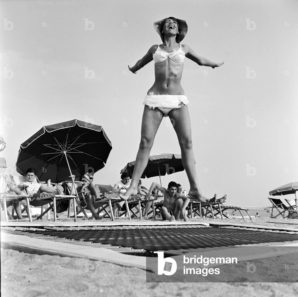 A model wearing bikini swimwear bounces on a trampoline on a beach in the French Riviera. 2nd August 1961 (b/w photo)