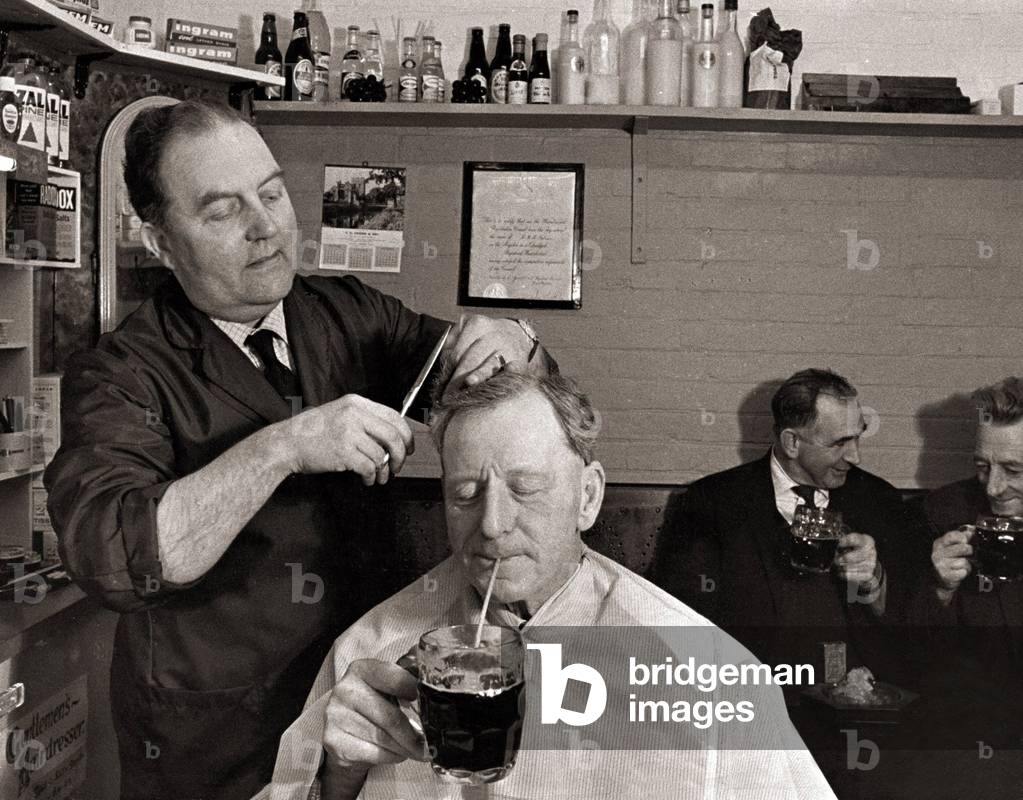 The landlord of the Railway Inn, Bernard Fabian cuts hair in between pulling pints in the little shop attached to the pub. When requested he can supply straws for customers like this gentleman who want to drink their beer while Bernard is cutting. November 1966 (b/w photo)