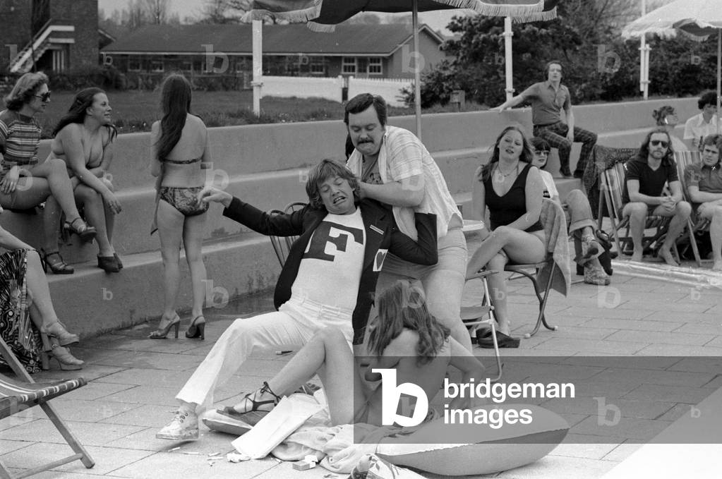 Robin Askwith and cast filming 'Confessions from a holiday camp' on Hayling Island. Robin Askwith removes bikini top from Janet Edis with husband Mike Savage watching who then throws him in the pool. 24th March 1977 (b/w photo)