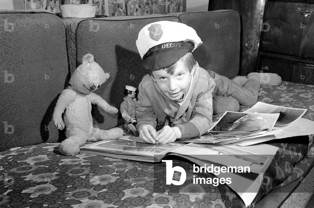 A little boy wearing a sailors cap as he hlies down on the sofa adding pictures of ships to his scrapbook