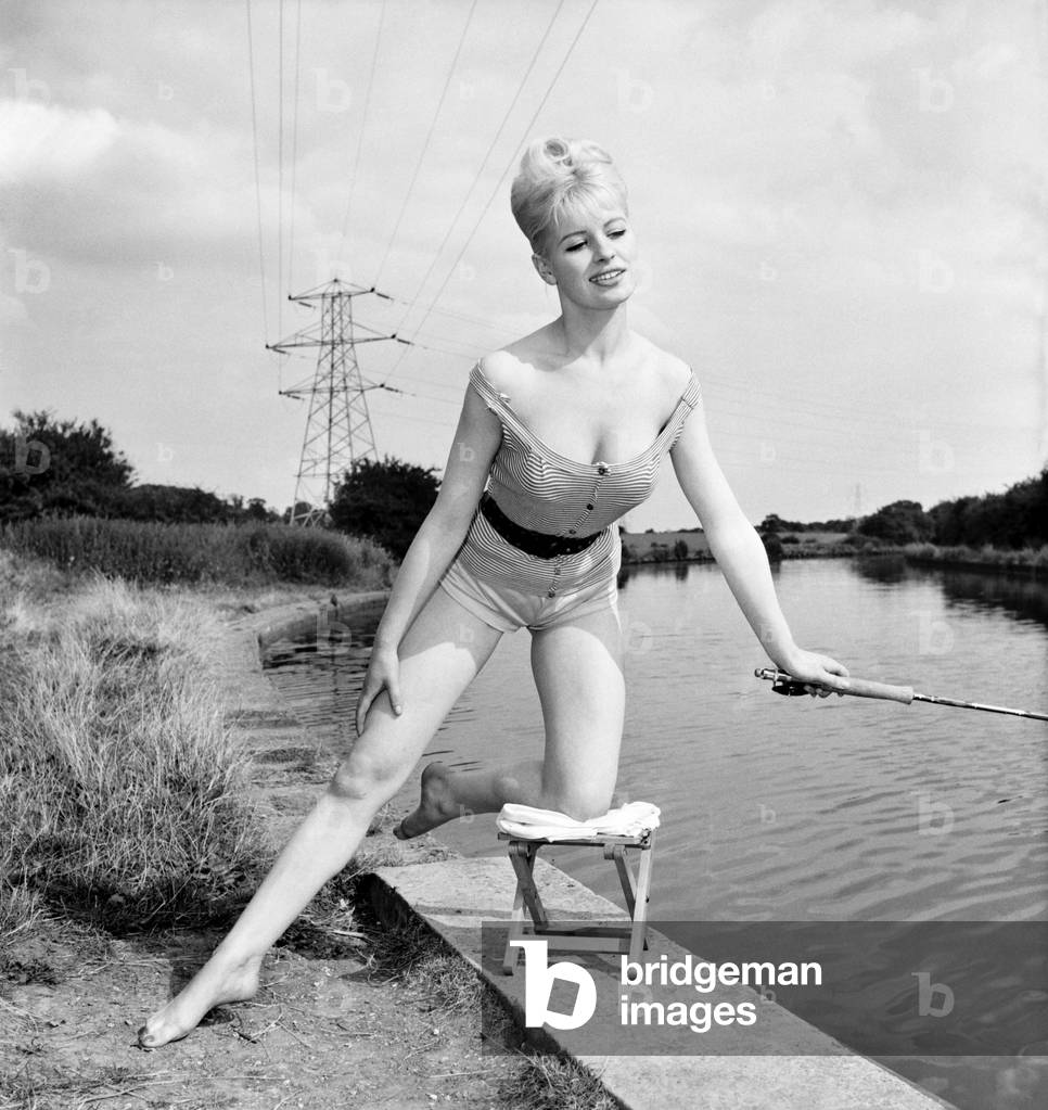 Beachwear model by the river bank fishing, August 1963 (b/w photo)