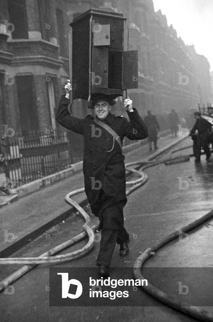 A cheery policeman with a cabinet balance on his head helps salvage furniture following a V2 attack on Woodbine, 3rd February 1945 (b/w photo)