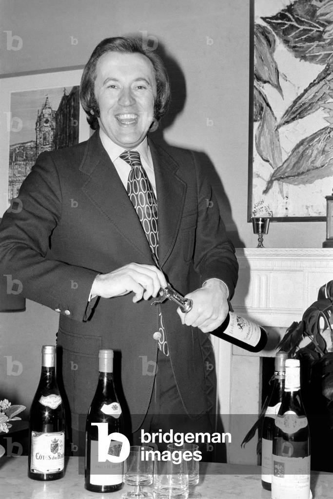 Mr. David Frost trying to open a bottle of wine at his home, March 1975 (b/w photo)