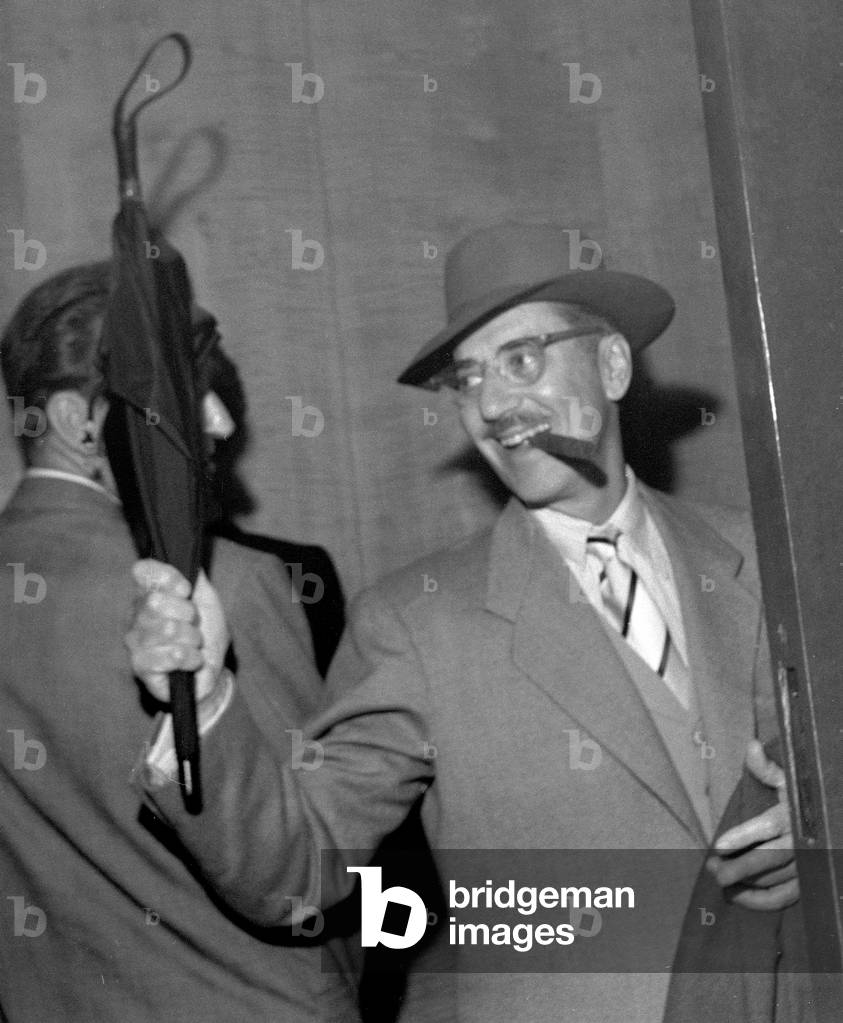 Comedian actor Groucho Marx, one of the famous Marx Brothers, enters the Savoy hotel in London, smoking a Cigar and waving an umbrella June 1954 (b/w photo)