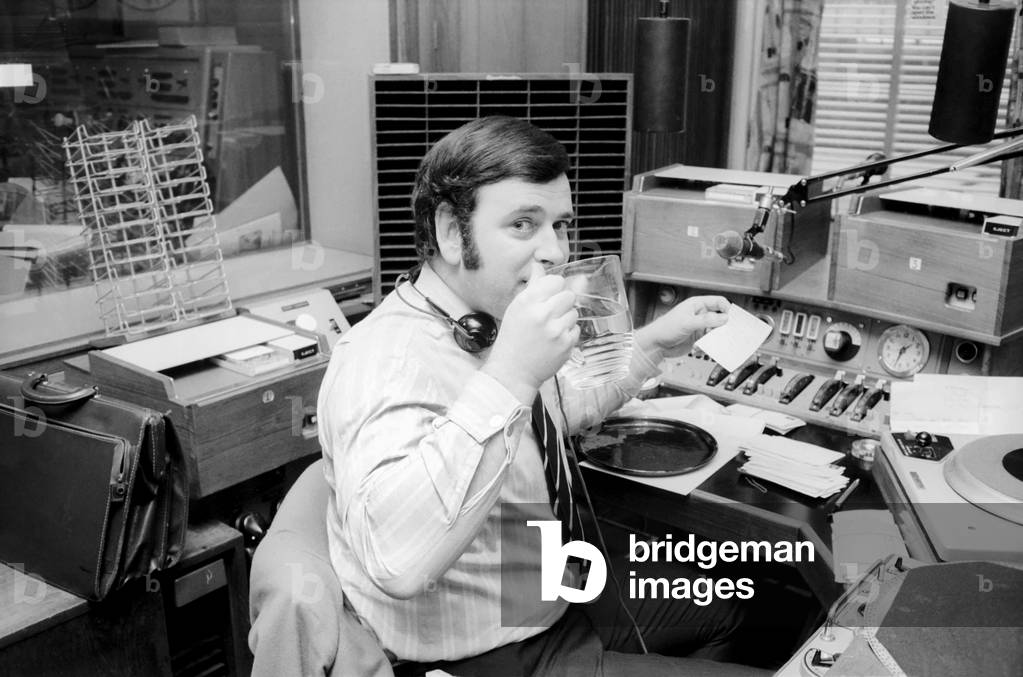 Disc Jockey Terry Wogan enjoys a large jug of water during broadcast of his afternoon radio show (2pm - 4, 15) on BBC Radio 1, January 1970 (b/w photo)