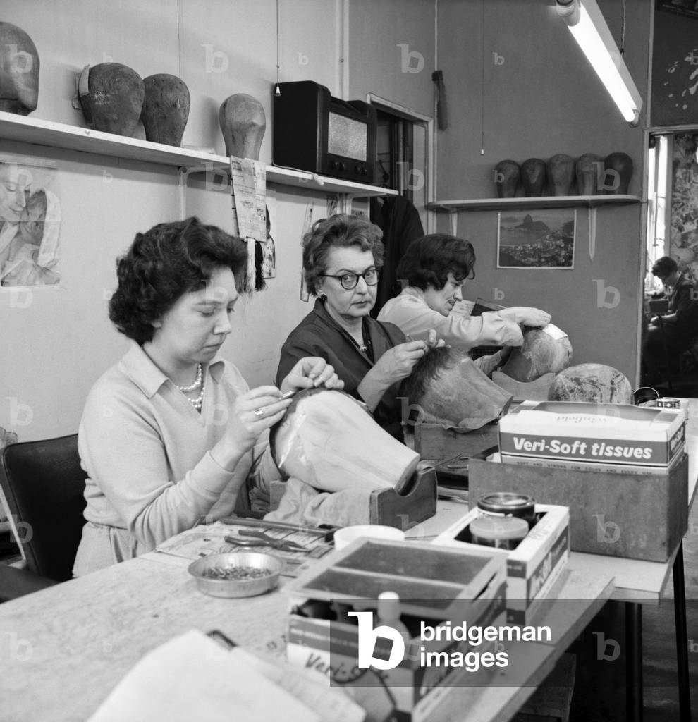 Wigmakers Workshop: Women, Making wigs whilst in another part of the workshop hair is selected for the next batch of wigs, 1963 (b/w photo)