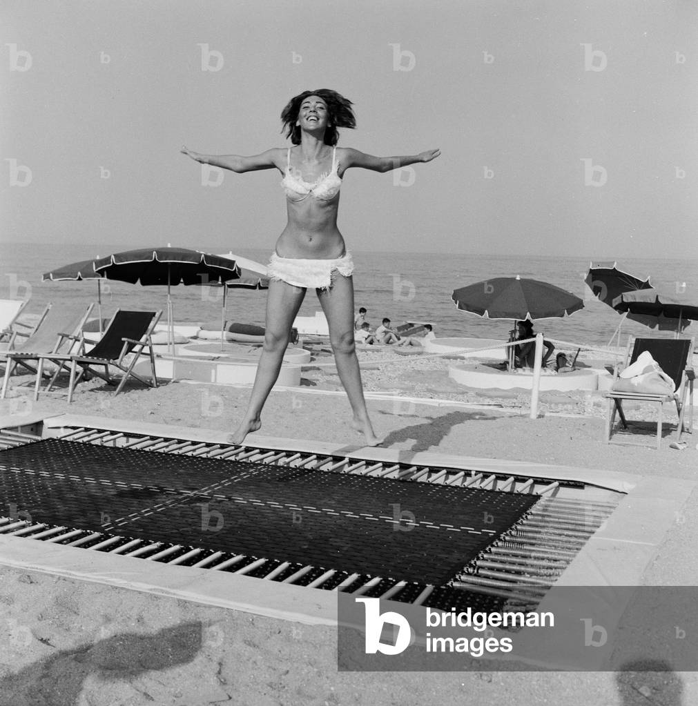 A model wearing bikini swimwear bounces on a trampoline on a beach in the French Riviera. 2nd August 1961 (b/w photo)