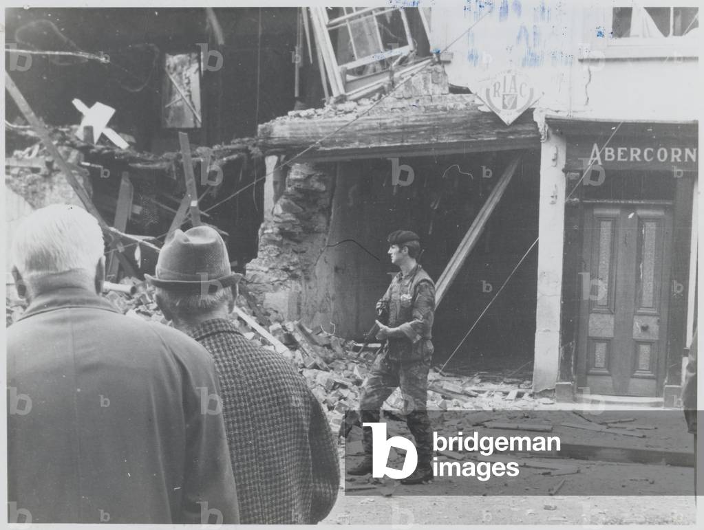 A soldier from the 1st Battalion The Royal Regiment of Fusiliers patrolling past onlookers and a building that has been blown up, Northern Ireland, c.1974 (b/w photo)