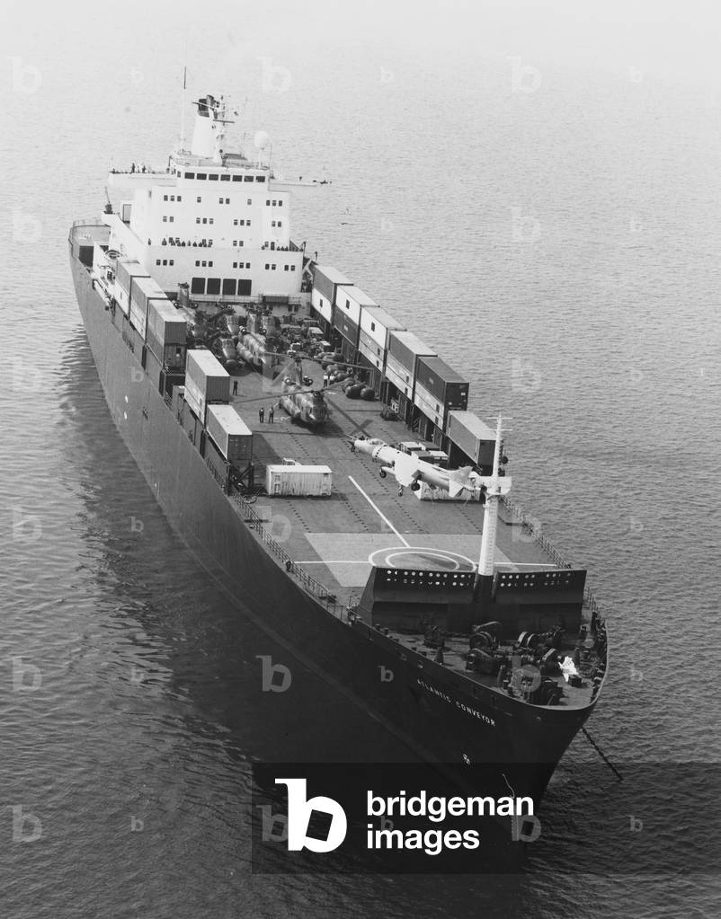 The ship Atlantic Conveyor with Chinook and Sea King helicopters loaded and a Sea Harrier hovering above the deck, South Atlantic, 1982 (b/w photo)
