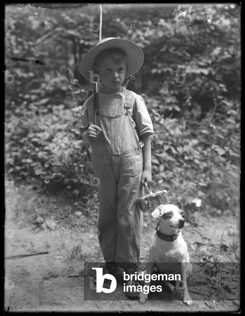 William Gray Hassler in straw hat and overalls, carrying fishing pole and basket, posed in the woods with a small dog (Bessie), August 20, 1911 (b/w photo)