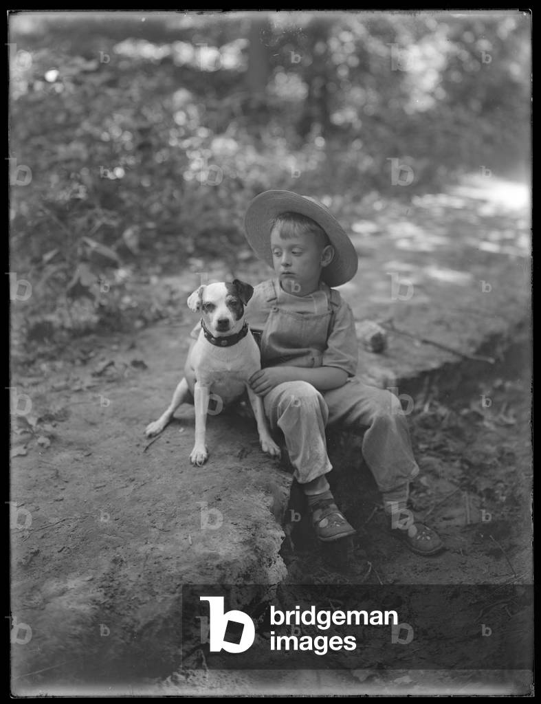 William Gray Hassler in straw hat and overalls, posed seated in the woods with a small dog (Bessie), August 20, 1911 (b/w photo)