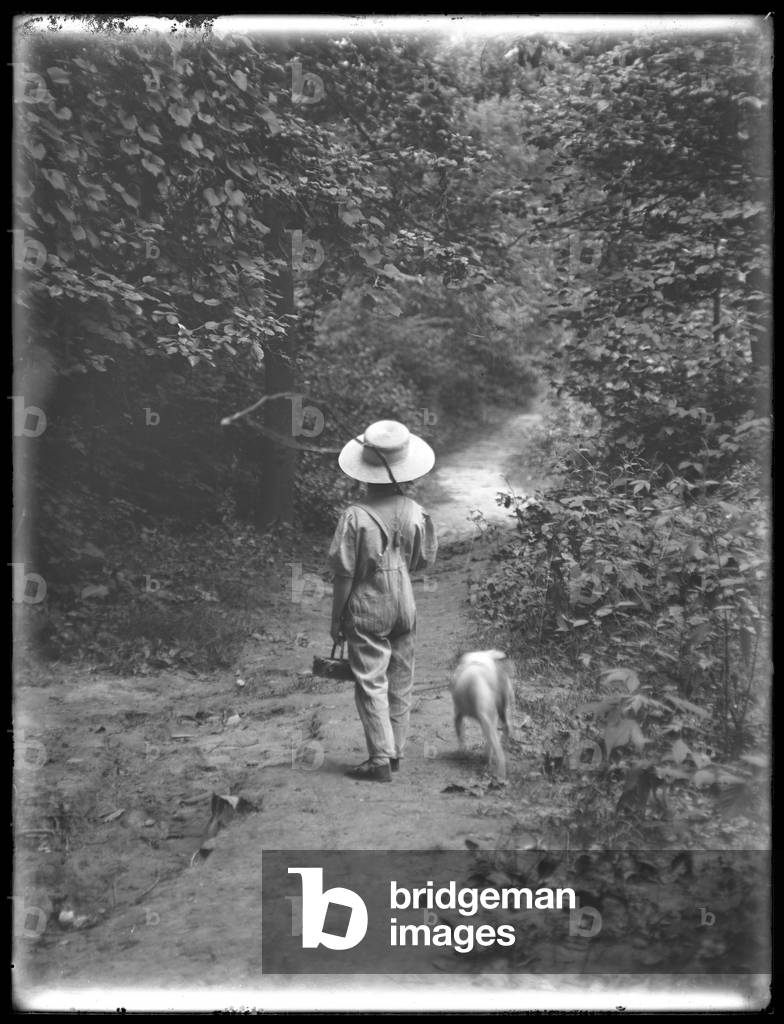 William Gray Hassler in straw hat and overalls, carrying fishing pole and basket, walking down a path in the woods with a small dog (Bessie), August 20, 1911 (b/w photo)
