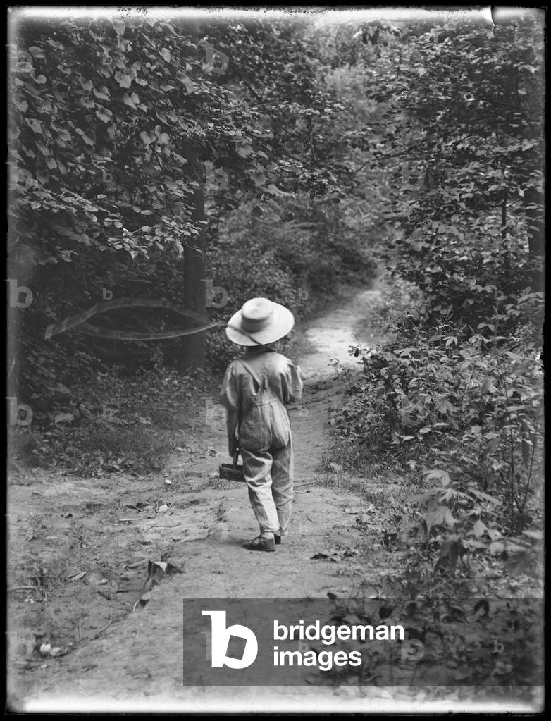 William Gray Hassler in straw hat and overalls, carrying fishing pole and basket, walking down a path in the woods alone, August 20, 1911 (b/w photo)