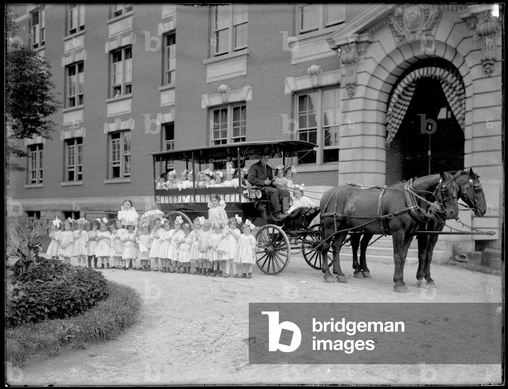 Small girls pose in and in front of a horse-drawn wagon, in front of an unidentified building, probably the Roman Catholic Orphan Asylum, Kingsbridge, Bronx, c.1910 (b/w photo)