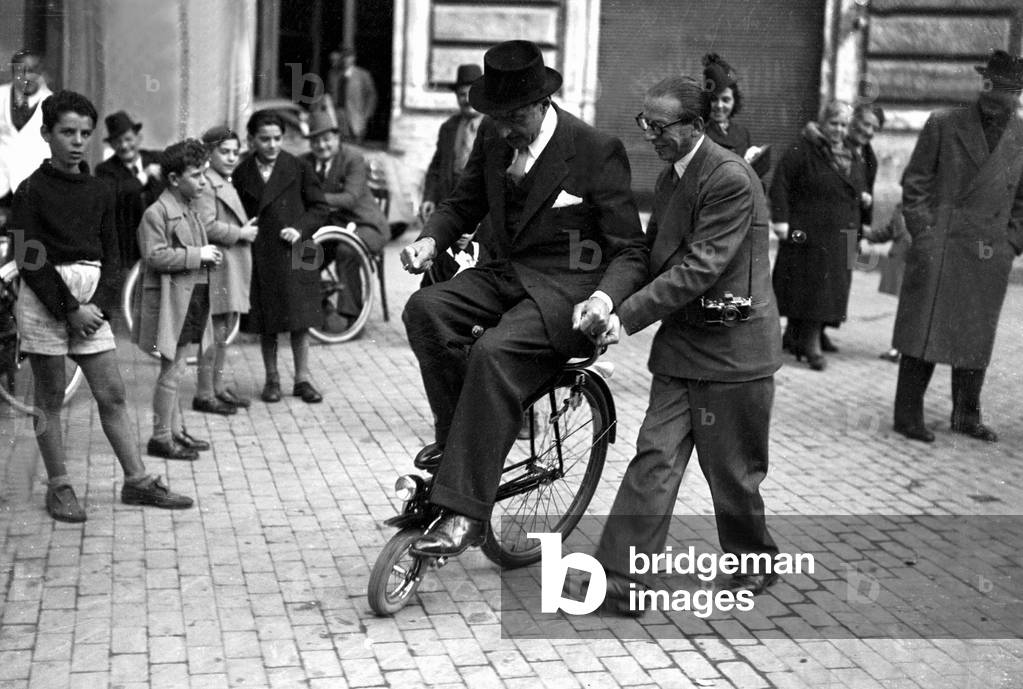Rome, 19/11/1939. Le poéte dialectal romain Trilussa ( Carlo Alberto Salustri) essaye le Velocino, petit vélo aux roues de dimensions différentes et au guidon placé à l'arrière . Le photographe Adolfo Porry Pastorel l'aide à l'utiliser. © Farabola/ Leemage