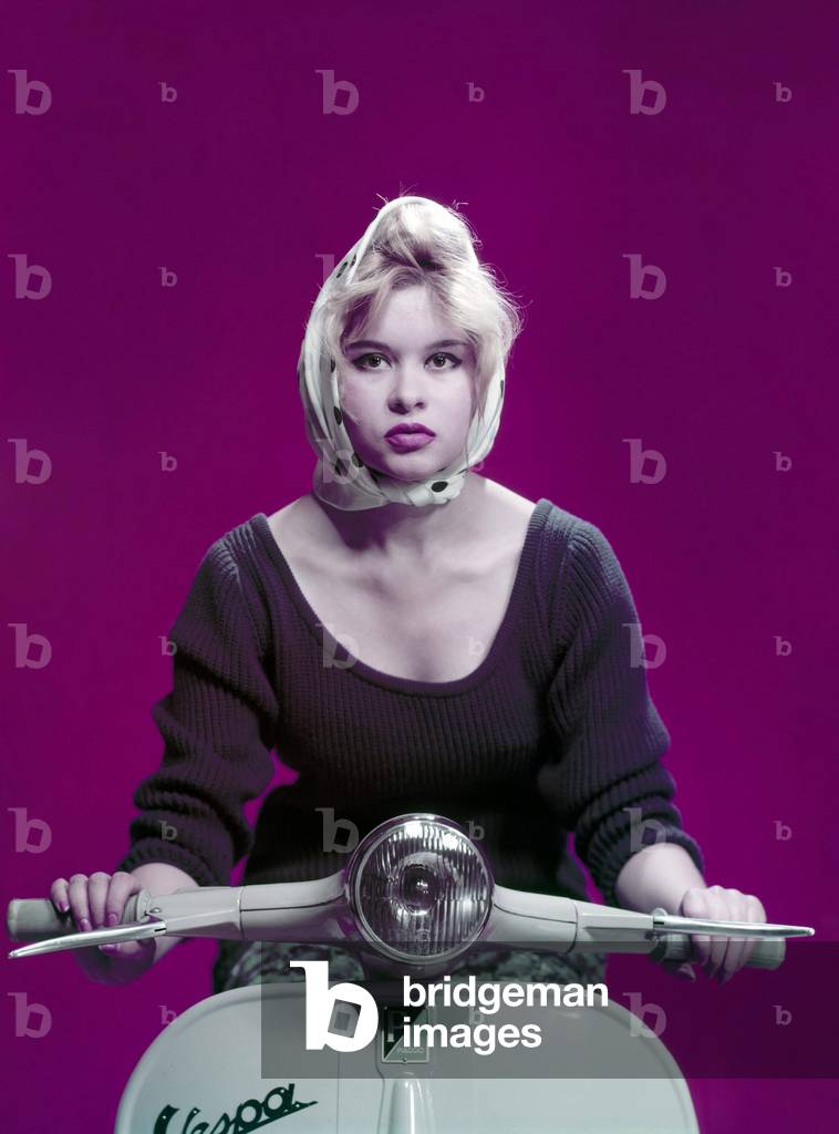 A young girl with a scarf in her hair poses at the wheel of a vespa, 1960 (photo)