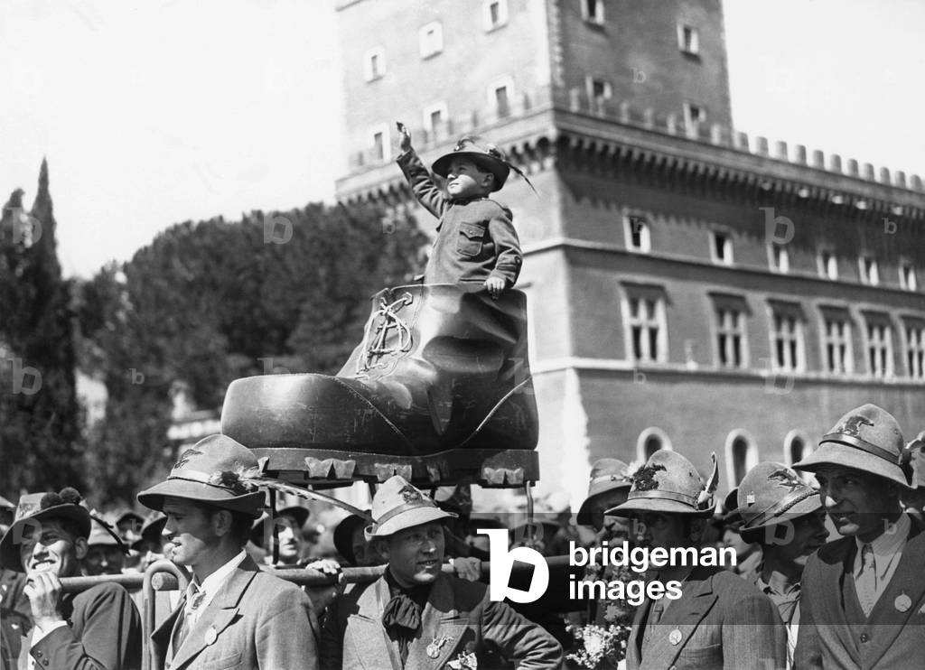 Parade of alpine hunters, carrying a child in uniform, in a large shoe, Rome, 1934 (b/w photo)