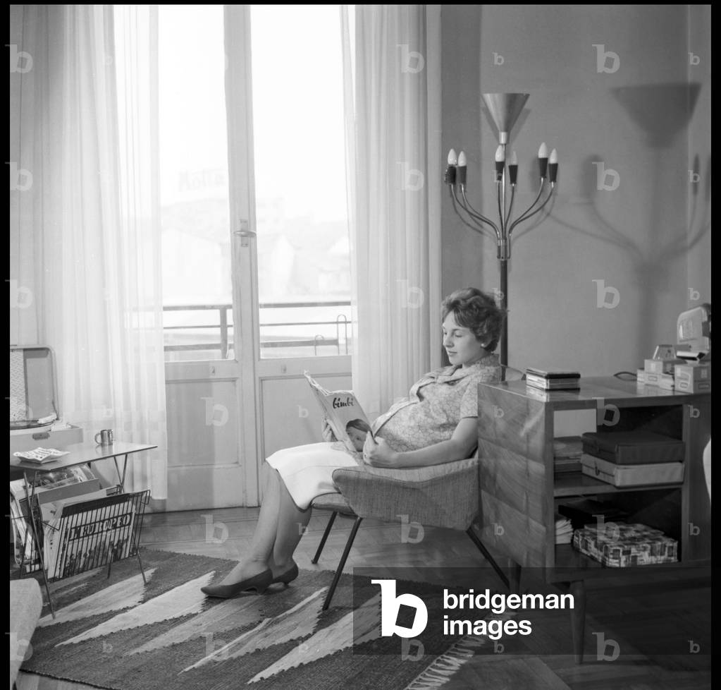 A pregnant woman reads a magazine for expectant mothers in her living room, Milan, 1959 (b/w photo)