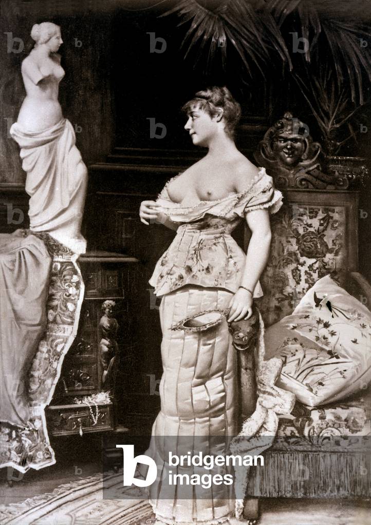 A young woman in a dress was photographed in a studio, innocently revealing her breasts in front of a statue of a nude woman inspired by antiquity.