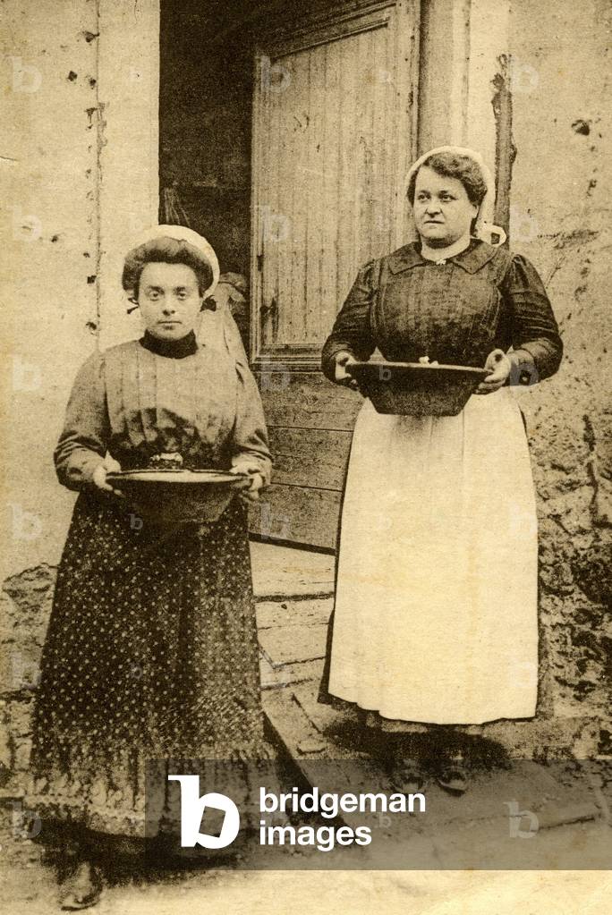 On the doorstep of the house, two villagers present the terrines they have just cooked. Photo at the beginning of the 20th century.