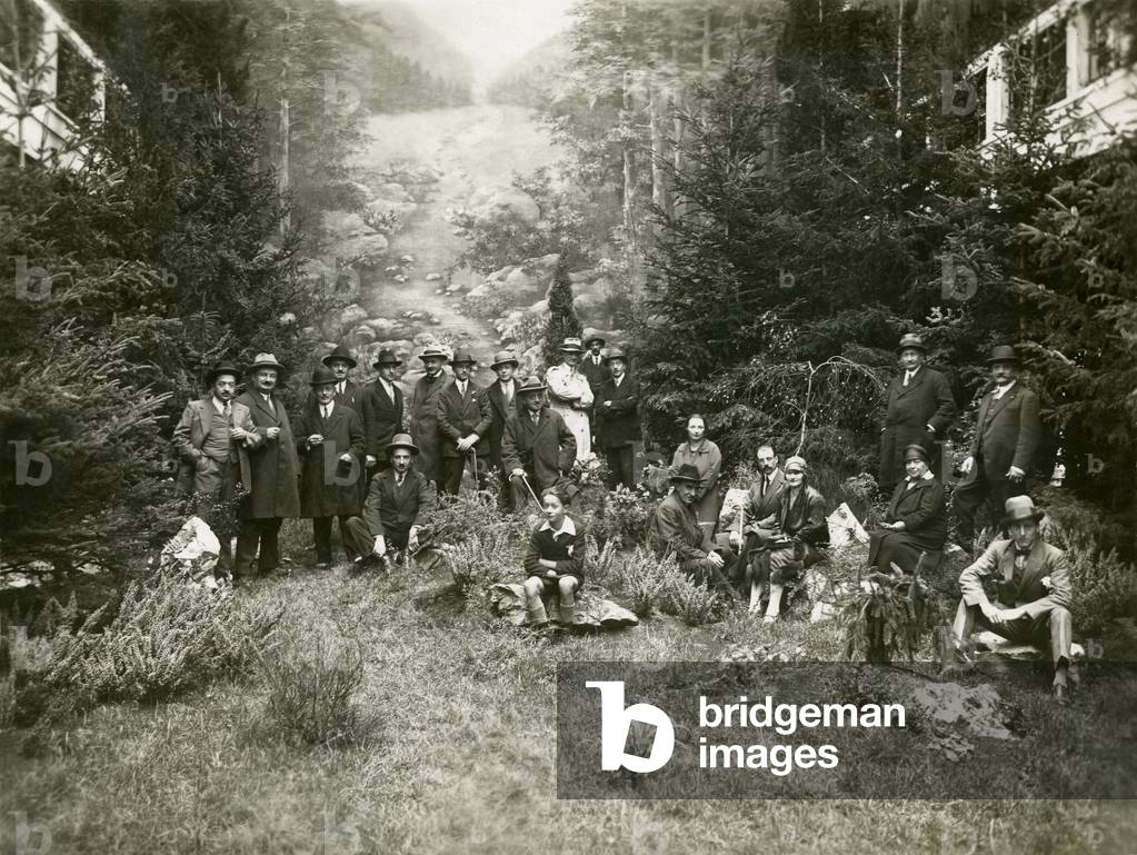 group of people posing for photography in a natural decor reconstructed inside a large exhibition hall - if the vegetation in the foreground and the trees on the coastline seem real natural, the background is clearly a painted background, representing a mountain stream. Anonymous print, ca. 1920-1930 -