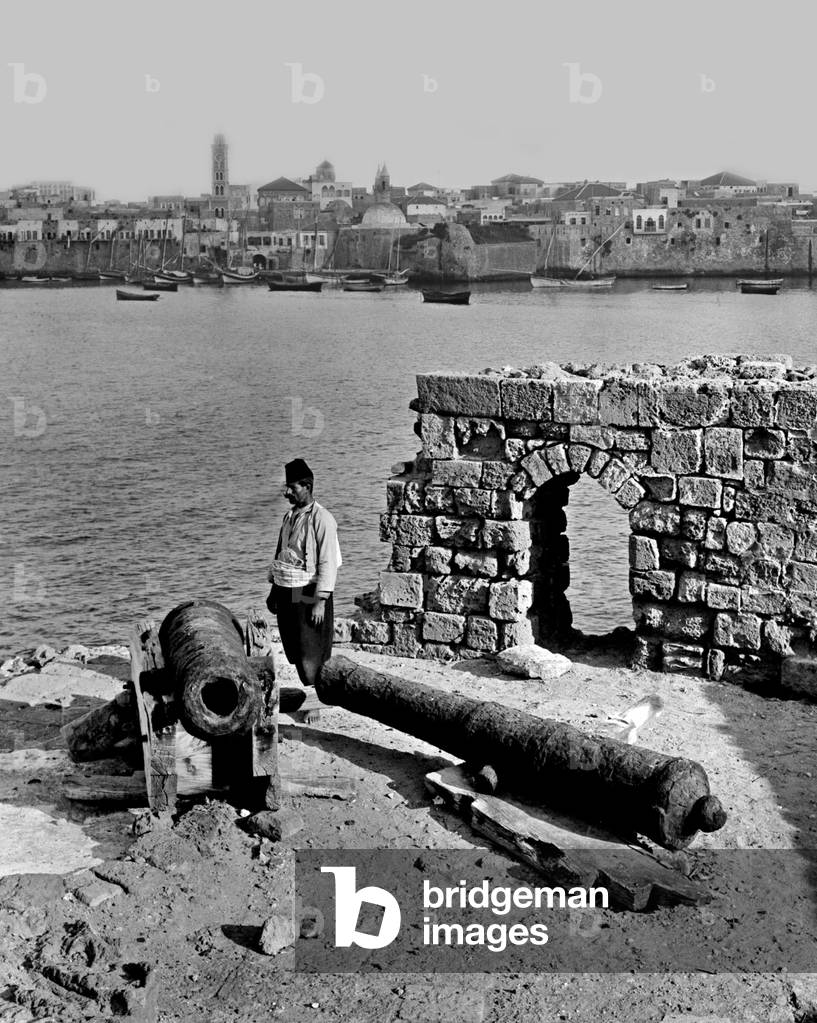 Palestine: A Palestinian man standing by an old canon at Acre, c. 1910