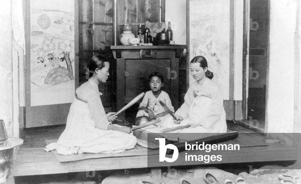 Korea: Two women using heavy pangmangi batons to smooth clothing, watched by a small girl. Early 20th century