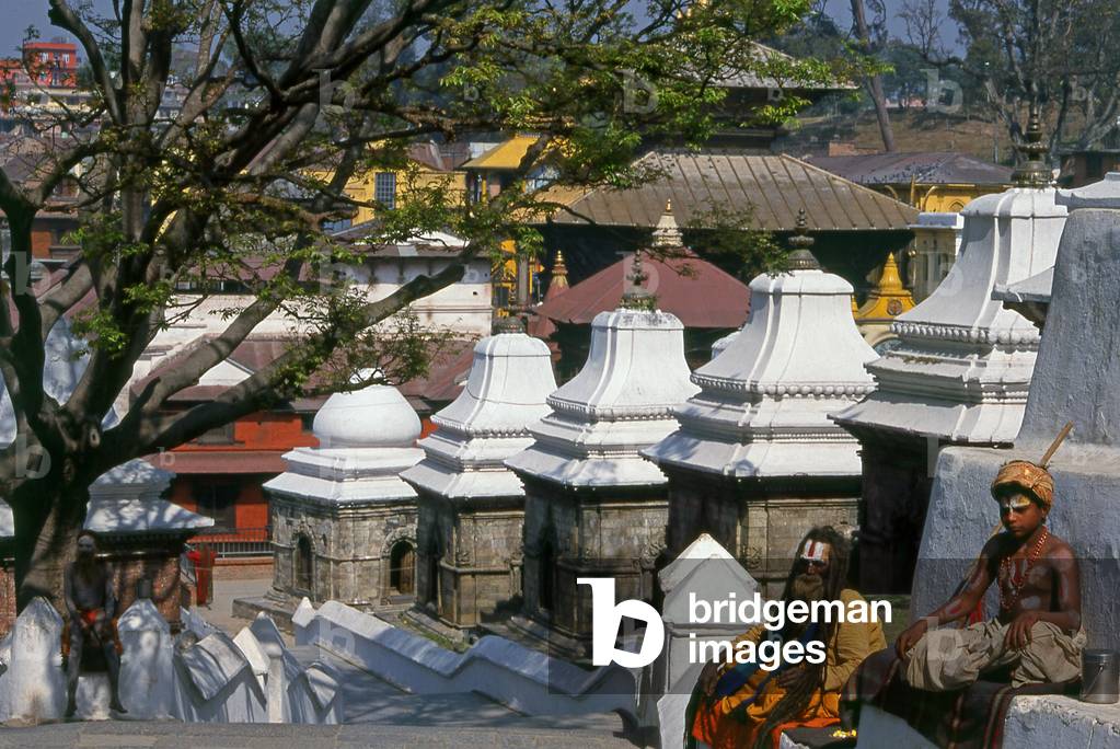Nepal: A sadhu (holy man) with a young disciple next to the chaityas (small Buddhist shrines) across the Bagmati River next to the Pashupatinath Temple complex, Kathmandu (1997) (photo)