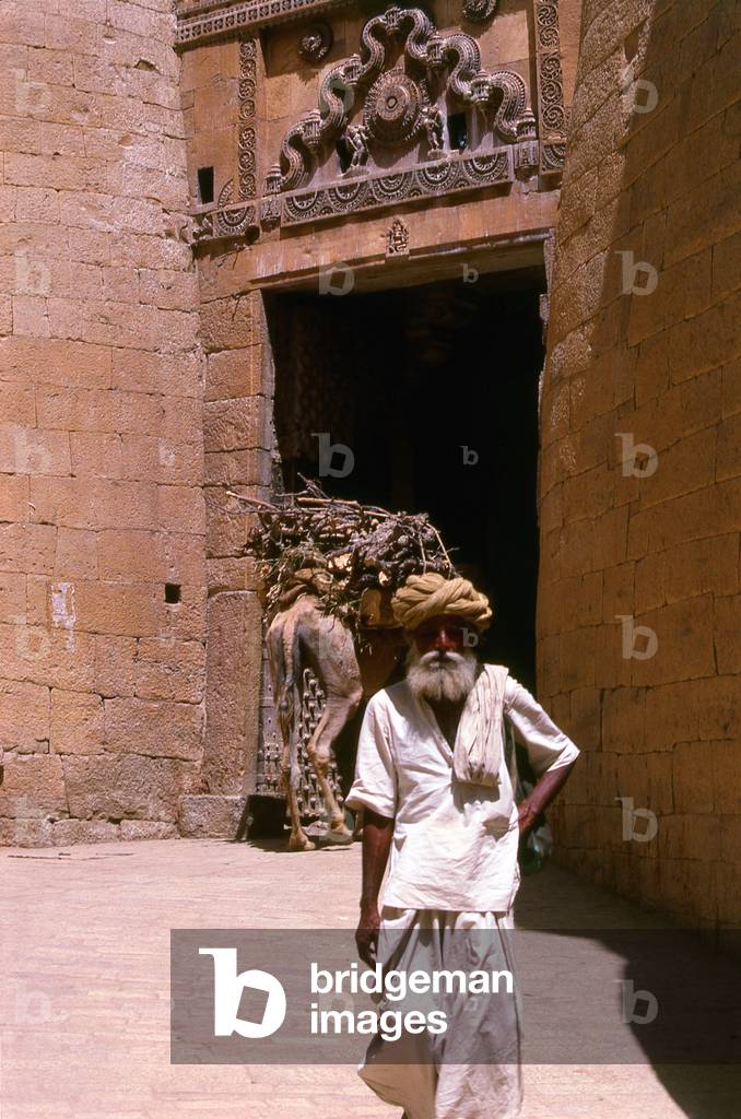 India: A heavily laden camel enters Surya Pol (Surya Gate) as a Rajasthani man exits, Jaiselmer, Rajasthan