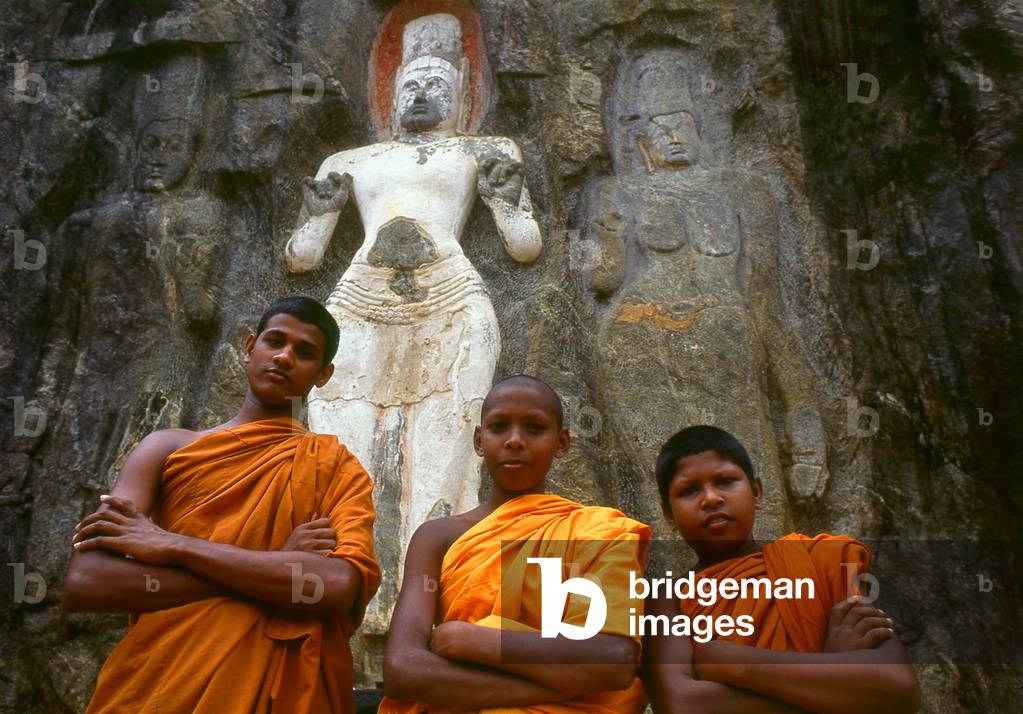 Sri Lanka: Young Buddhist monks in front of 1000 year old carved stone figures representing the Avalokitesvara (centre) and his consort Tara (right), and their son Prince Sudana, Buduruvagala (photo)