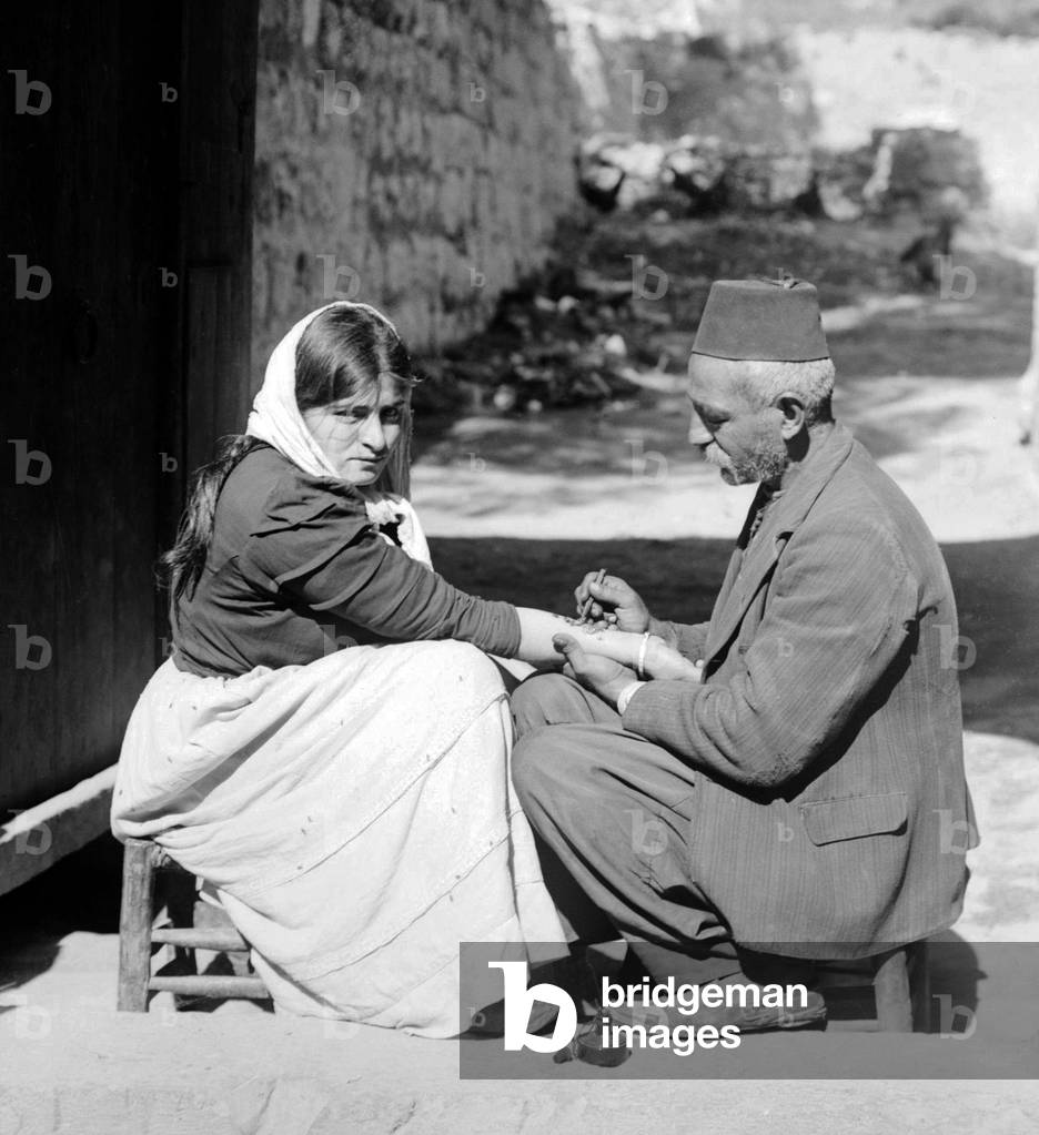 Palestine: Palestinian man tattooing a religious charm on a Christian woman's forearm, c. 1920