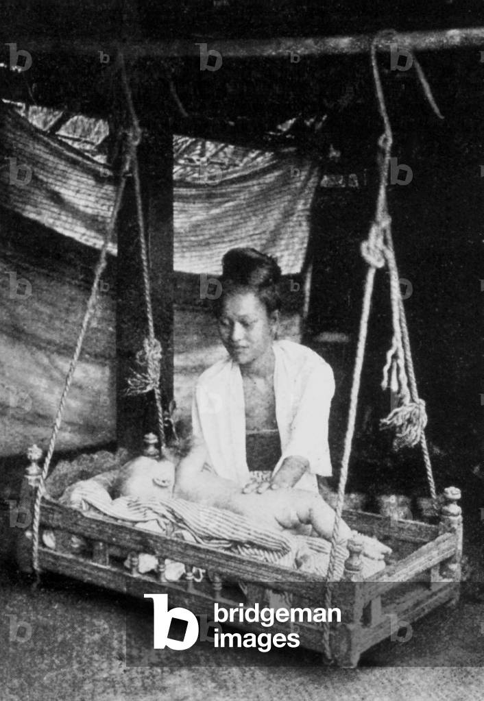 Burma: A Burmese mother cares for her child who lies in a swinging wooden cradle (Burmese: 'saungban'), c.1892-96.