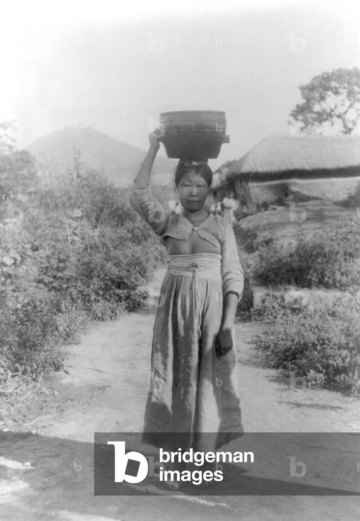 Korea: A peasant woman carrying a pot of water on her head in a rural scene near Seoul, early 20th century