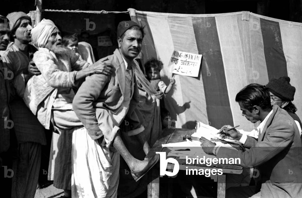 India: First Lok Sabha general election, Delhi, January 1952. A blind old man being carried towards a polling booth by his son at a polling station near the Jama Masjid (Friday Mosque) in Old Delhi