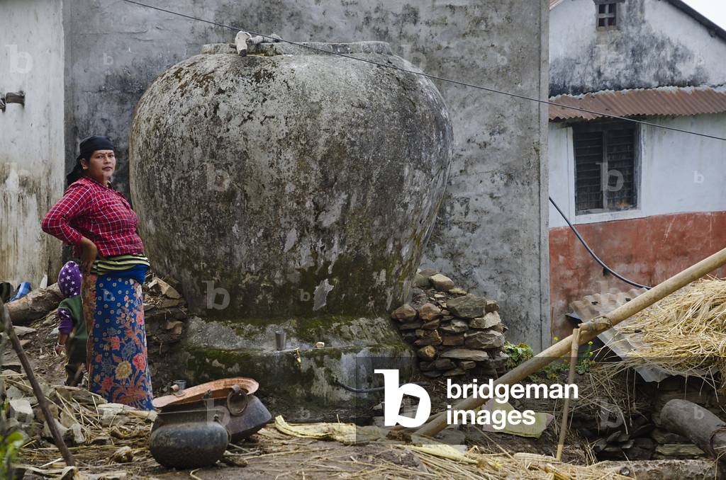 Nepali woman standing next to urn-shaped traditional water storage tank, 
Majhthana, near Pokhara, Nepal (photo)
