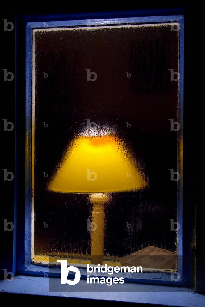 Warm glow of table lamp in dark room behind rainy window, Gower, South Wales, UK (photo)