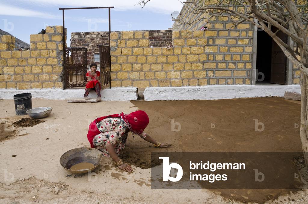 Woman applying cow dung mixed with water & earth to patio floor outside her home in a desert hamlet, Thar Desert, Rajasthan, India (photo)