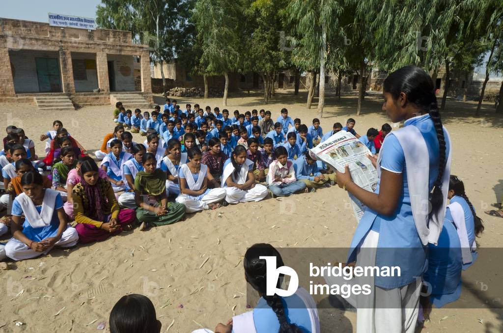 Schoolgirl reading newspaper article to pupils sitting in schoolyard,
Gagadi (aka. Gagari) village, Rajasthan, India (photo)