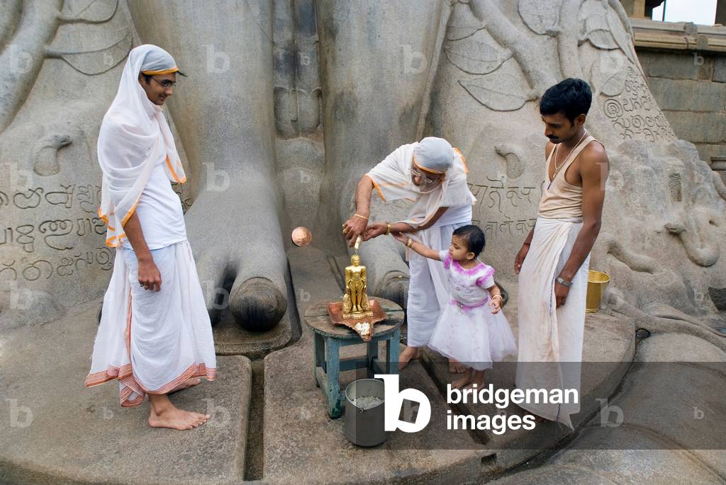 Jainism: Devotees initiating little girl into ritual worship at feet of 10th century Gomateshwara sculpture, Indragiri Hill, Sravanabelagola (one of prime Jain pilgrimage centres), Karnataka, India (photo)